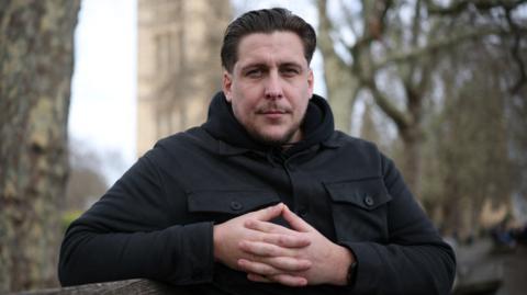The image shows a man wearing a black jacket while interlocking his fingers together. Behind him trees and a building can be seen.