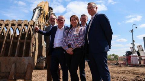 Four people stood in front a construction site with a large JCB digger 