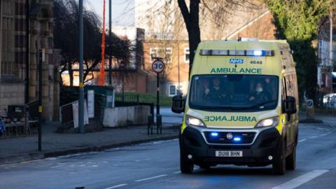 An ambulance with its blue lights on is driving down a road, towards the camera. In the background buildings can be seen, along with a 30 mph speed limit sign