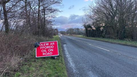 Arwydd 'Olew ar y Ffordd' ar ran o'r A5025 ger Pentraeth, oriau wedi gwrthdrawiad yn yr ardal yn cynnwys dau gar