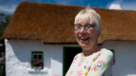 Margaret Gallagher pictured smiling outside her thatched cottage in July 2019.  She has short grey hair and glasses and is wearing a yellow floral top.