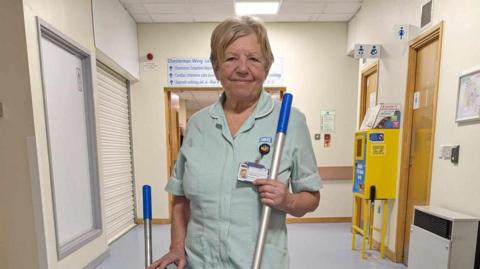An older lady with short blonde hair in scrubs, standing in a hospital hallway, holding a mop handle.