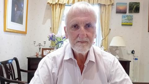 A man with white hair and white facial hair. He is wearing a white shirt with blue stripes. Behind him is a window with yellow curtains, and several pictures hung on the wall