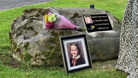 A woman with a black and white printed dress, which is the only part of her in the picture, is looking at a memorial to Arlene Arkinson. There is a plaque to her on a stone and a picture of her as a child in a frame next to it. There are also some flowers on the stone.