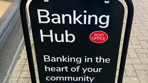 A black sign on a street with white writing that says "banking hub, banking in the heart of your community".