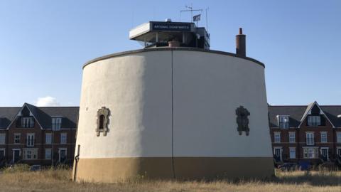 The round grey exterior of Martello Tower P in Felixstowe featuring small windows reinforced with bricks and a National Coastwatch lookout on top 