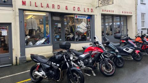 Millards bike shop from the exterior. A creamy yellow building with motorbikes in front and a red sign which says Millards & Co Ltd.