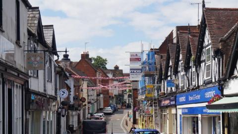 General view of Droitwich Spa High Street. A view down a narrow high street lined with a mix of historic black-and-white timber-framed buildings and modern shopfronts. Several cars are parked along the road, while decorative bunting is strung across the street overhead. Scaffolding can be seen on one building in the distance, and a number of businesses, including shops and takeaways, are visible beneath a partly cloudy sky.
