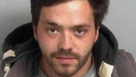 Charles Benson-Clark wearing a grey crew-neck shirt layered under a dark jacket, seated against a plain light grey background. He has black hair.