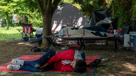 A fully clothed man lying down on a blanket in front of several tents in a park-like setting