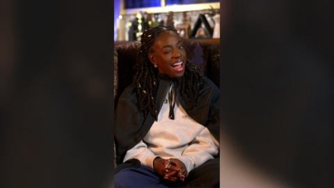 Cat Burns winks while sat in an armchair wearing a dark green cloak over a grey sweater. She has long black braids