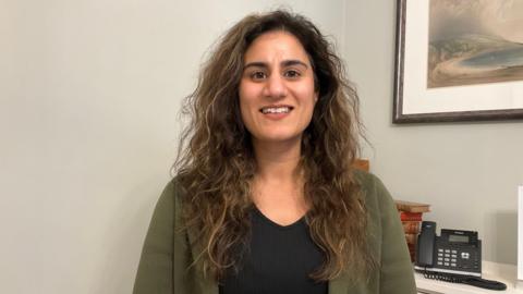 A woman in her 20s with shoulder-length wavy hair stands indoors against a light-coloured wall, wearing a dark top and green cardigan. Behind her is a white cabinet with a desk phone and stacked folders, and a framed coastal-style artwork hanging on the wall.