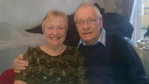 Mavis and Dennis are sitting together, and Dennis has his arm around his wife. He had short white hair and glasses. Mavis has short blonde hair.