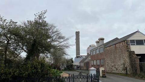 A chimney out of the power station. In the foreground is a tree and a building made of bricks with a series of windows. 