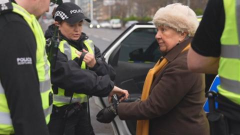 A woman, wearing a brown jacket and cream-coloured puffy hat, is arrested by a female officer next to a police car. Two other officer stand on either side of them in the foreground. 