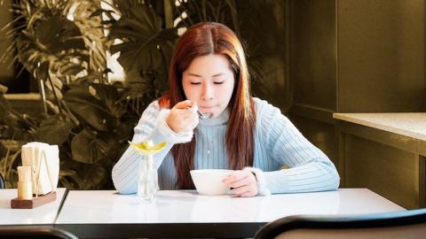 A woman sits at a table and eats from a small white bowl.