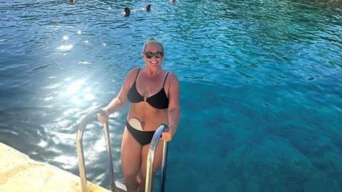 Lindsay Bowers is smiling at the camera, wearing dark sunglasses and a black bikini. She is holding on to step handles as she climbs into a swimming pool.
