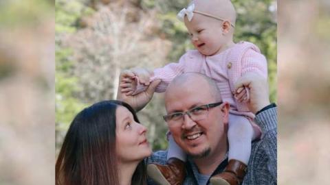 Chris Wade with a bald head and glasses wearing a light blue striped jumper. He is smiling. He has a little girl, Layla, in a pink outfit on top of his shoulders. His wife Josianne Charlebois has brown hair and is wearing a flowery black top. She is looking sideways at her daughter on her husband's shoulders. 