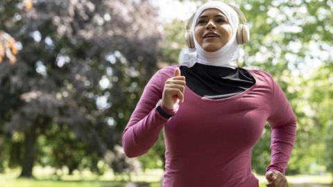 A woman in a white headscarf and a pink long-sleeved t-shirt runs through a leafy park. She is also wearing white headphones.