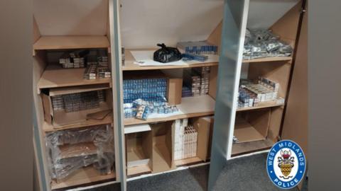 Boxes of counterfeit cigarettes sit on shelves. A West Midlands Police logo is in the bottom right-hand corner of the picture.