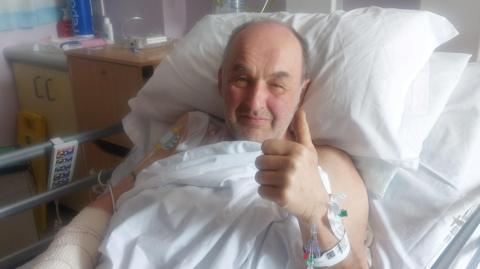 Joe Hughes, lying in a hospital bed, holding up his left hand, with medical equipment in his arms, sitting on white sheets and pillows, and smiling at the camera. He has short grey hair and a stubbly beard. There are two hospital cabinets to the right. 