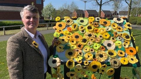 Lindsey Barrow, with short grey hair, a white shirt and brown dogtooth jacket, stands in the sunshine next to the information board, which is covered in knitted and crocheted yellow poppies
