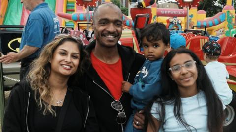 A woman, man and two children are smiling at the camera. The man is holding a small boy in his arms and a young girl is standing beside them.