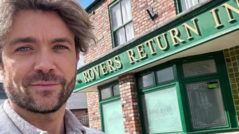 Emrhys Coopers stands in front of Coronation Street's Rovers Return Inn. The pub has a red brick exterior with a green facade. The letters 'Rovers Return Inn' are written in gold.