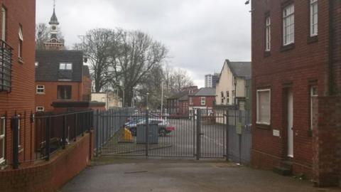 A view of the existing grey metal car park gates to the building which are located between two red brick buildings 