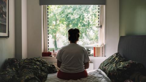 Rear view of a young woman sitting an unmade bed, looking out of the window at trees outside