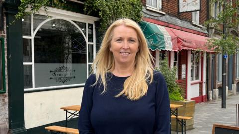 A woman with blonde hair smiling standing outside a pub with an Italian restaurant in the background