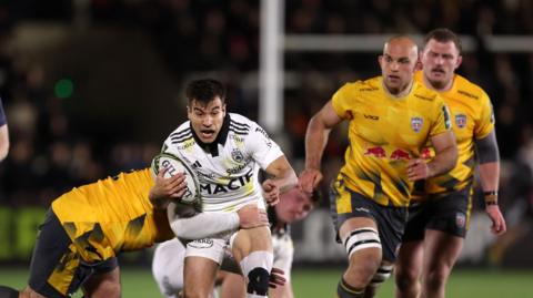 La Rochelle scrum-half Thomas Berjon carrying the ball but is tackled by a Newcastle Red Bulls player whose face is obscured, with two other Red Bulls players watching on the right