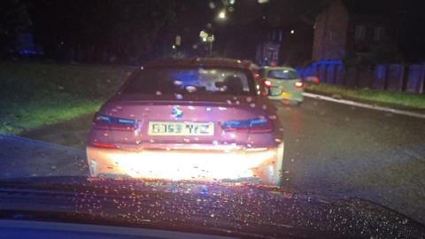 The back of a red car with the number plate blurred. The car is on the side of the road at night.