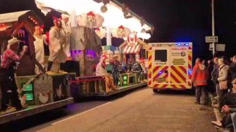 An ambulance driving along the road between a crowd of onlookers, and a line of carts. There is also a carnival marshal wearing an orange hi-vis vest following the ambulance.