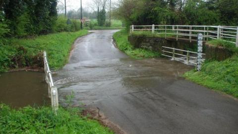 A ford with a river running over the road. Next to the ford is a bridge. There are white railings on both of them, trees on both sides of the road, and a road junction in the distance.
