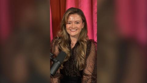 A woman with long brown hair, in front of a pink curtain, smiling to camera.