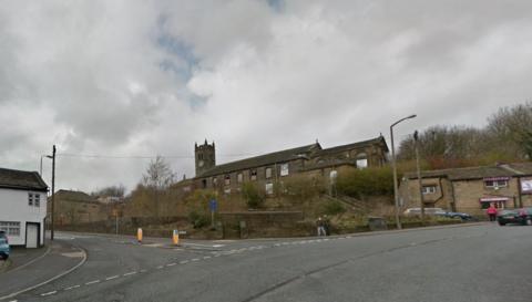 The Keighley Road looking left towards Raw Lane. An old white building with black window and door frame is on the left, with a parked car. A crossing island is in the centre of road. An old stone church is on the hill with two pedestrians nearby. A pedestrian and two cars are close to a stone building on the right with purple signs.