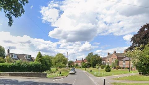 A junction with a road curving away into the distance. Grassed areas on either side of the road, with trees and large village houses set back from the road. The sky is blue with some clouds.
