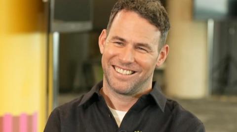 Mark Cavendish is smiling, he has short dark hair, and is wearing a black shirt, with a white t-shirt underneath.