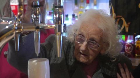 Hilda Hilton pulls a pint at the Shield and Dagger pub. She is wearing a grey coat and glasses. She has white hair. There are drinks behind her. She has a pint in front of her.