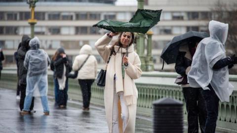 A woman wearing a cream coat and scarf struggles with her green umbrella due the strong winds on the Westminster Bridge in London. 