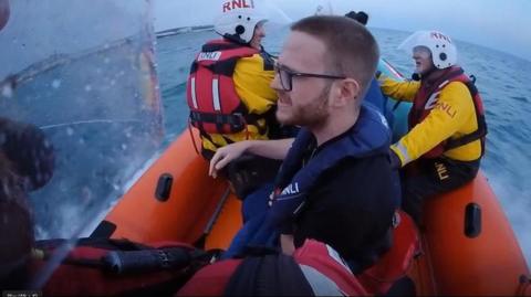 A man with short hair and glasses sits in a lifeboat at sea as he is rescued by RNLI volunteers.  
