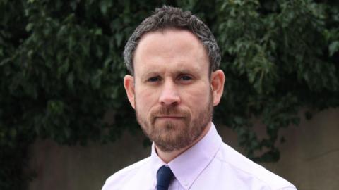 Andy Symonds in a pale pink shirt and navy blue tie looking directly at the camera. 