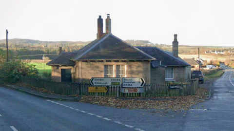 Two roads meet on a corner. A house in situated on the corner, with a sign pointing left for Gordon and Edinburgh and right for Stichill and Greenlaw. It is a countryside setting, with green fields and bushes in the background