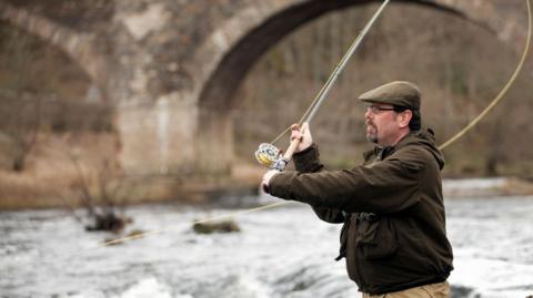 A man wearing a flat cap and brown jacket casts a fishing rod