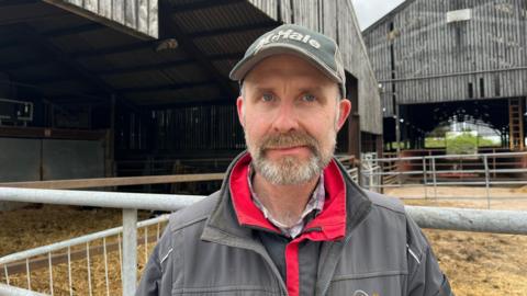 Photograph of Chatham looking into the camera with a neutral expression. He has a short, dark grey beard. He is wearing a red and black waterproof jacket with a grey overall over the top. Chatham is also wearing a dark green cap with 'McHale' written on it in white lettering. He is stood in front of two huge barns, clad in corrugated iron. There are metal gates and fences in front of the entrances, with straw strewn across the floors. 