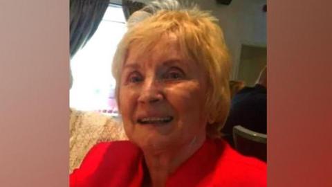 Jean Hindmarch. She has short blonde hair and is smiling at a camera. She is sitting on a sofa and is wearing a red blazer.