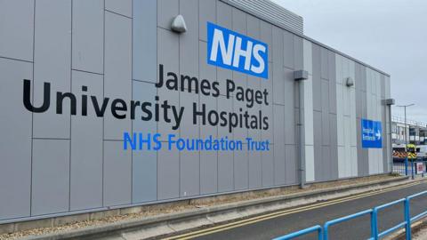 A grey wall of a hospital which says NHS James Paget University Hospitals and in front of it a road. Beyond it to the far right can be seen two ambulances