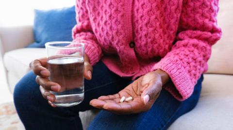 Woman wearing bright pink cardigan and dark blue jeans hold two pills in one hand and a glass of water in the other