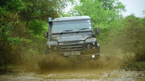 A Land Rover vehicle driving towards the camera through a muddy puddle with vegetation and trees on either side.